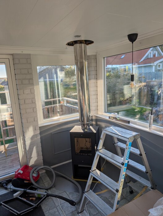 Installationen av en vedspis i ett nyrenoverat rum, med stege och dammsugare bredvid.
