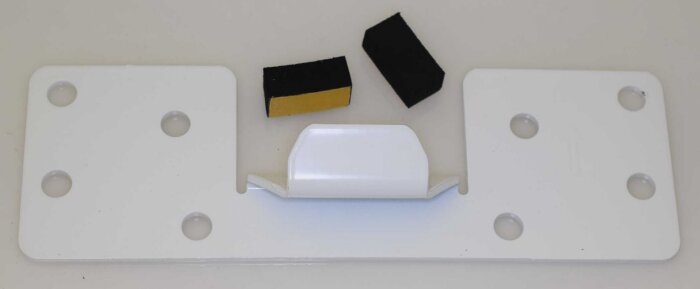 Vit monteringsfäste med fyra hål, en mittstång och två små svarta och gula skumdelar ovanpå, för installation.