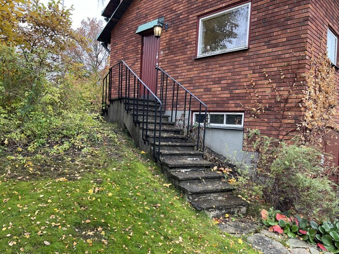 Betongtrappa med smidesräcke vid tegelvilla; entré med dörr ovanför trappan, omgiven av buskar och träd.