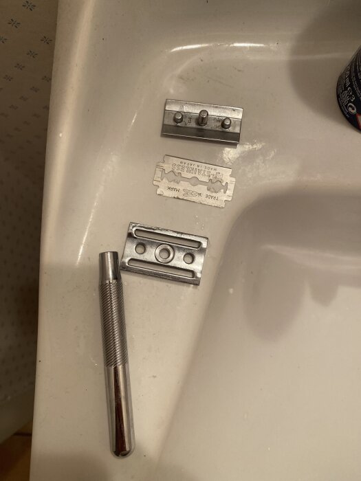 En isärskruvad safety razor med löstagbart blad, handtag och huvuddelar ligger på ett handfat. Bladet har en enkel egg på varje sida.