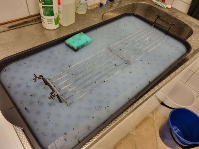 Grillgaller som rengörs i en skobricka fylld med vatten på en diskbänk; diskmedel och rengöringssvampar finns också i bilden.