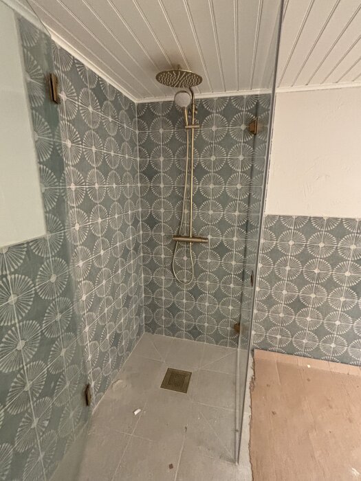 Dusch med mönstrade kakelväggar och regndusch; ej färdigställd utan dörr och fläkt.