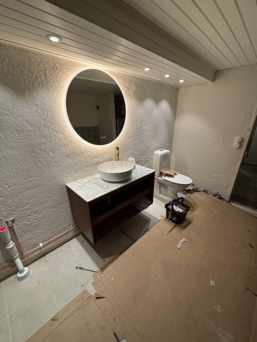 Ett nästan färdigt badrum med runt spegel, handfat och toalett, förberett för dörr och fläkt.