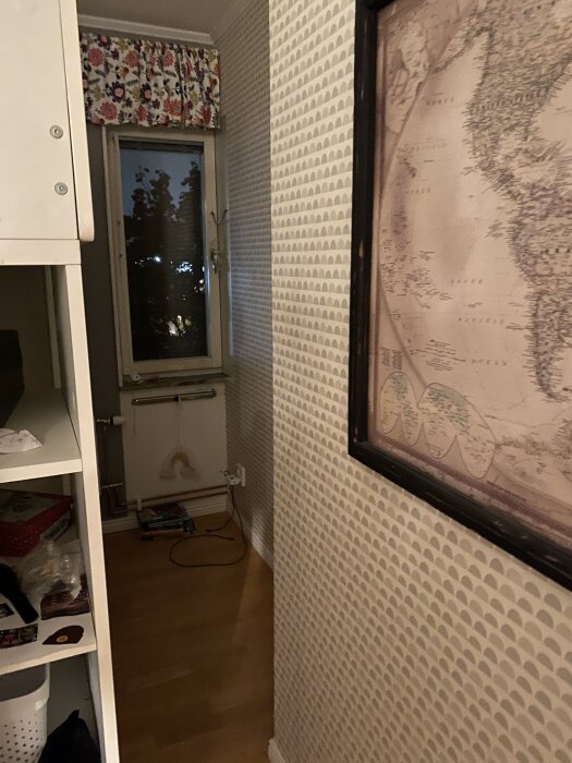 En smal hall med trägolv, tapetmönstrade väggar, ett långsmalt fönster med blommigt draperi och en karta i ram på väggen.