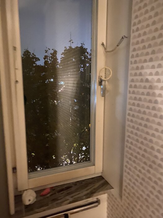 Fönster med mörker utanför, synligt träd, inomhusvägg med mönster, laminatskiva och fönsterhantag med snöre.