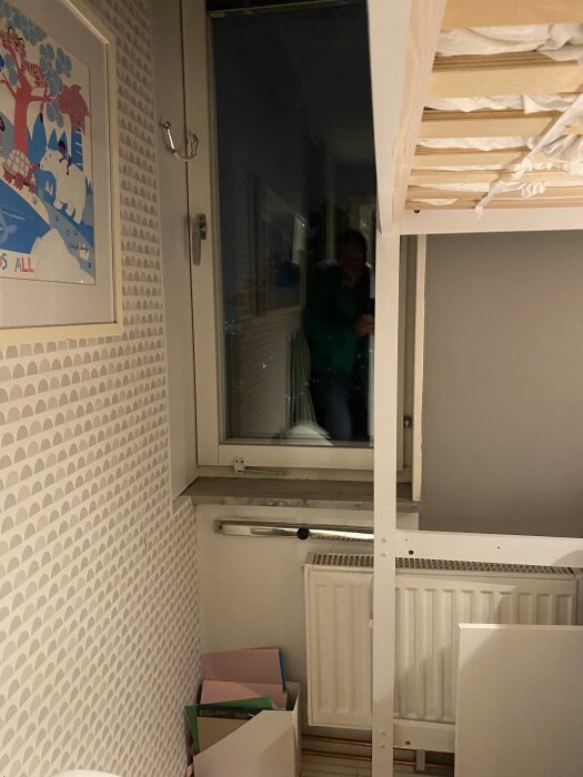 Rum med fönster, radiator, loftsäng och en tavla på väggen. En låda med papper under fönsterkarmen. Reflektion i fönstret.