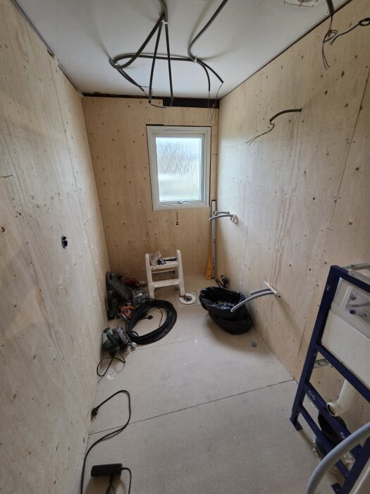 Rum under renovering med gipsväggar, ett fönster utan karm och diverse byggmaterial och verktyg på golvet.