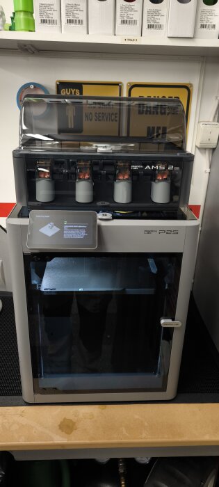 3D-skrivare med display och filamenthållare, placerad på ett träbord i verkstadsmiljö. Bakgrund med hylla och skyltar.