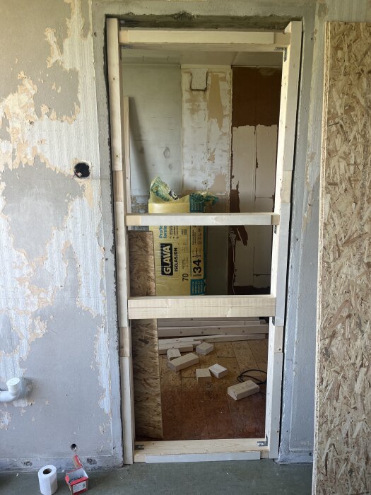 Öppning i betongvägg med träreglar förberett för igenbyggnad, OSB-skiva på sidan. Spår av renovering med ojämnt puts och tapetrester.