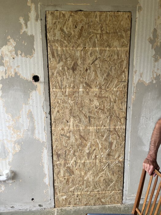 Dörröppning igensatt med OSB-skiva infälld i en grå betongvägg med avskavd yta, och en plastdetalj synlig i nederkant. Tänkt för renovering.