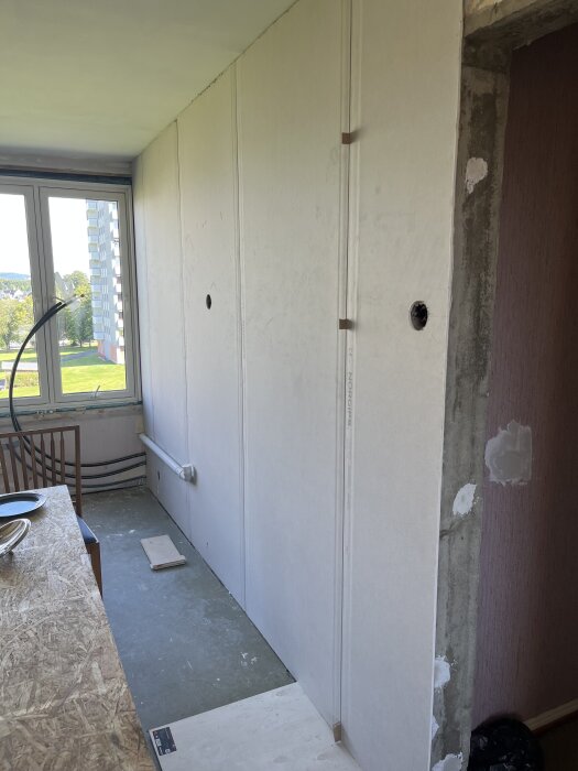 Gipsskivor på en vägg i en lägenhet under renovering, reglar och OSB i dörröppning, utsikt genom fönster mot grönska utanför, golvbrädor på betonggolv.