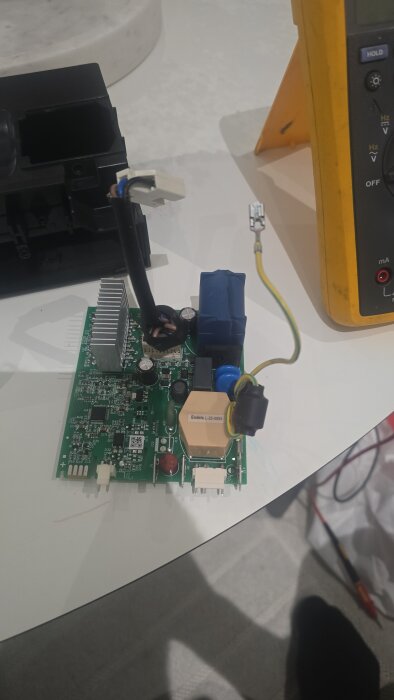 Grönt styrkort för kompressor med anslutna kablar på ett bord, delvis synlig multimeter i bakgrunden.