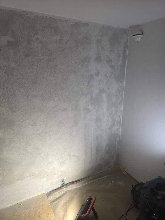 Vägg med pågående spackling, belyst med arbetslampa, sladd synlig längs golvet, i rum under renovering.