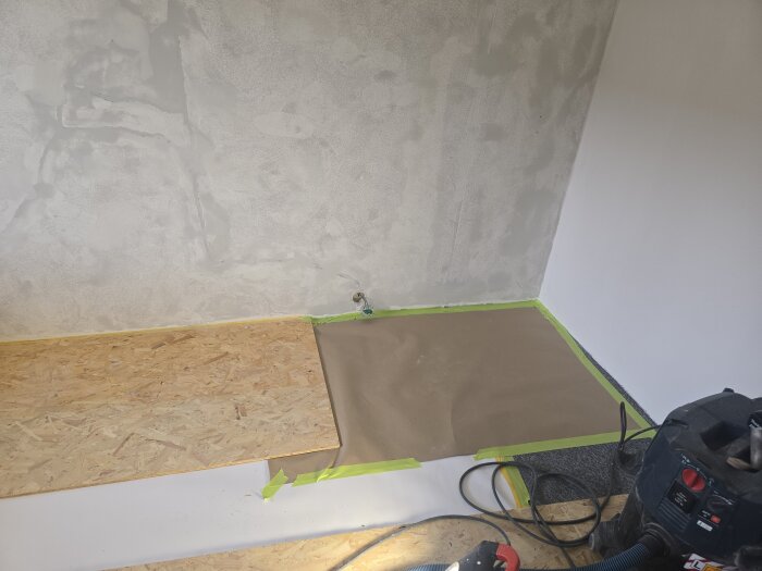 Vägg med spackel och grundfärg, golvet skyddat med OSB-skivor och papper, delvis täckt med maskeringstejp, dammsugare synlig.