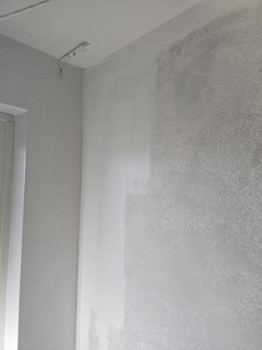 Bilden visar en vägg med pågående målningsarbeten. Ena sidan är jämn, medan den andra har fläckar av grundfärg och spackel.