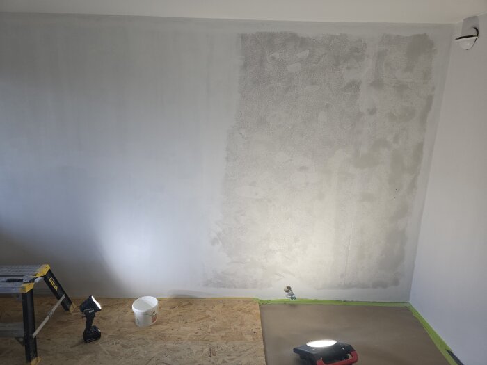Vägg under reparation i barnrum; halvt spacklad och grundmålad. Stege, verktyg och ett skyddstäckt golv synligt.