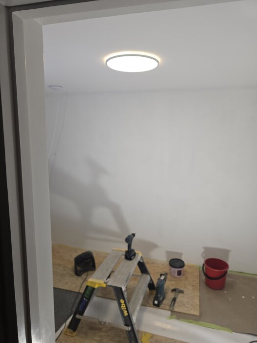 Rum under renovering med taklampa installerad, stege, verktyg och färgburk på golvet, förberett för målning och arbete.