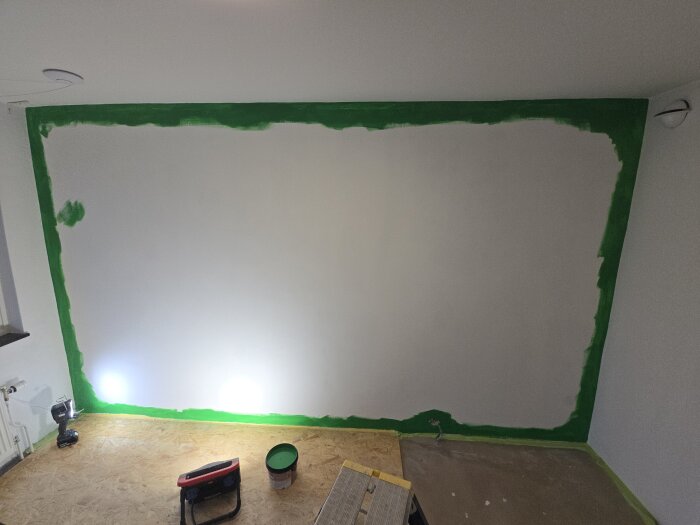 Grönmålad vägg i barnrum med ojämn täckning, golv av OSB-skivor, belysning på golvet och målarutrustning synlig.