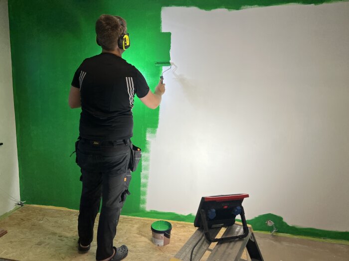 Person målar en vit vägg grön med roller, står på pall. Grön färgburk och strålkastare på golvet.
