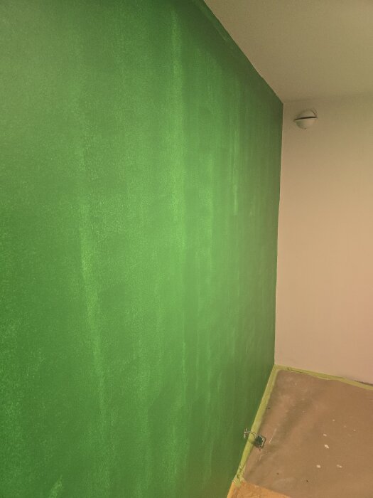Grönmålad vägg med ojämn yta, taklist synlig ovan och golv täckt med skyddspapper.
