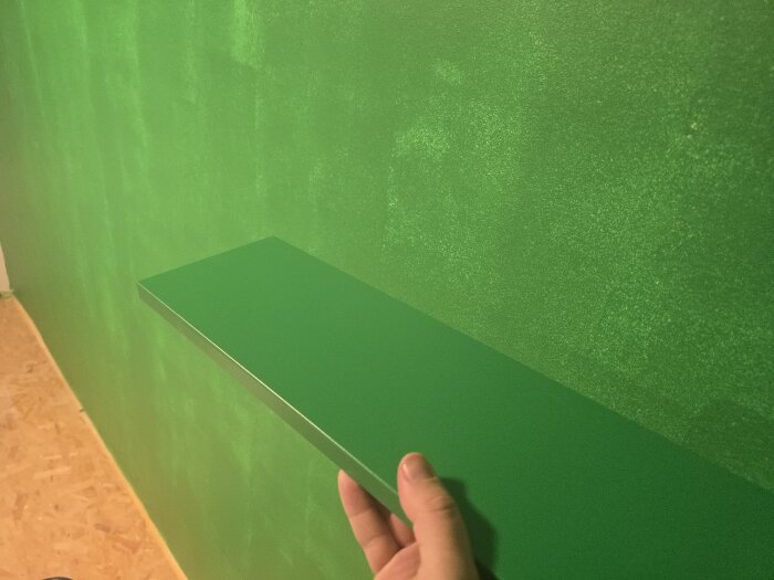 En grön hylla hålls upp mot en nymålad gröngränt vägg, golvet syns delvis.