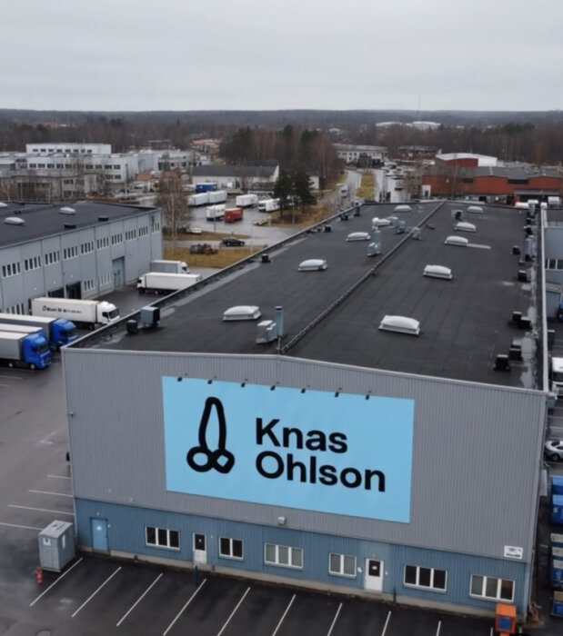 En industribyggnad med "Knas Ohlson" på fasaden. Flera lastbilar är parkerade runt byggnaden, och landskap i bakgrunden.