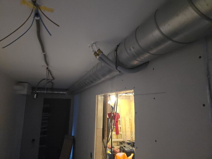 Byggrörelse med synliga ventilationsrör i taket och elektriska ledningar. Ett intilliggande rum syns genom en öppning.