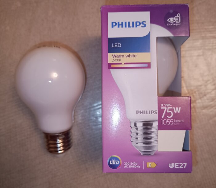 Philips LED-lampa 8.5W 2700K CRI 80 och dess förpackning, E27 sockel, varmvit ljus, 1055 lumen, ligger på trägolv.