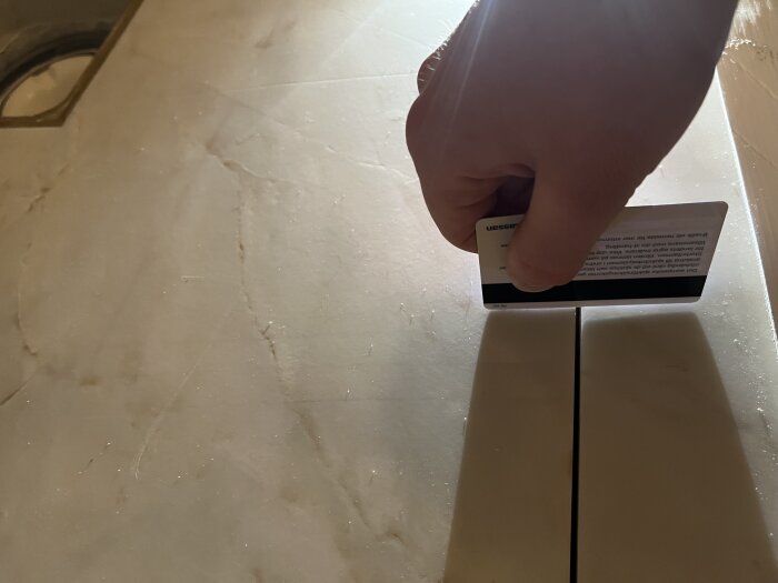 Hand håller ett bankkort mot en kakelplatta, visar fogens bredd. Belysning från ovan och kortet syns delvis. Tester av fogens jämnhet i ett badrum.