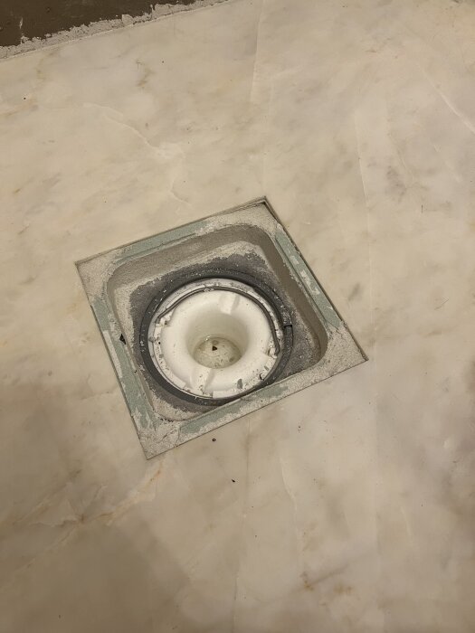 Golvbrunn i badrum med tecken på slitage och behov av slipning för nivåskillnad och fall till dusch. Kant och fog runt brunnen synliga.