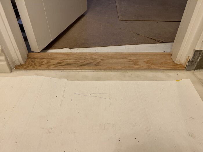 Tröskel av trä i dörröppning med synlig ojämn fogning mot ljusare golv, indikerar byggproblem och behov av åtgärd.