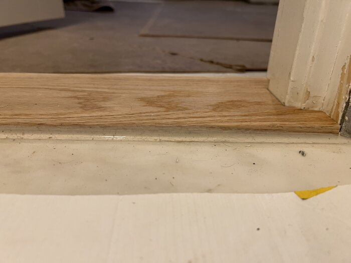 Tröskel med dåligt fogad kant, nära vit vägg. Golv och byggmaterial syns i bakgrunden. Bild från byggprojekt.