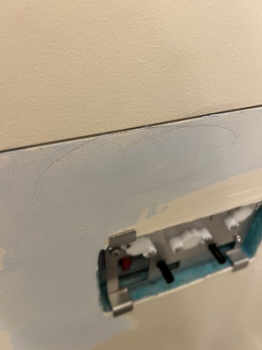 Närbild av vägg med oregelbunden yta och spackelmärken, samt ett infällt väggfäste med synliga beslag, troligtvis del av renoveringsprojekt.