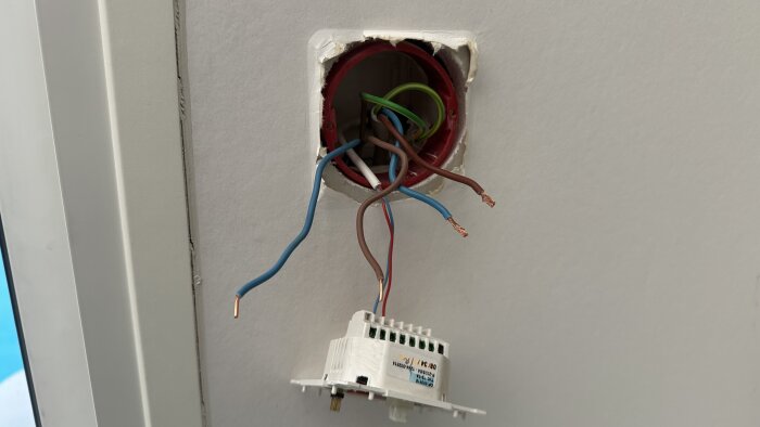 Elektrisk kopplingsdosa med frilagda kablar, några blå och bruna, och en vit komponentenhet som hänger från väggen.