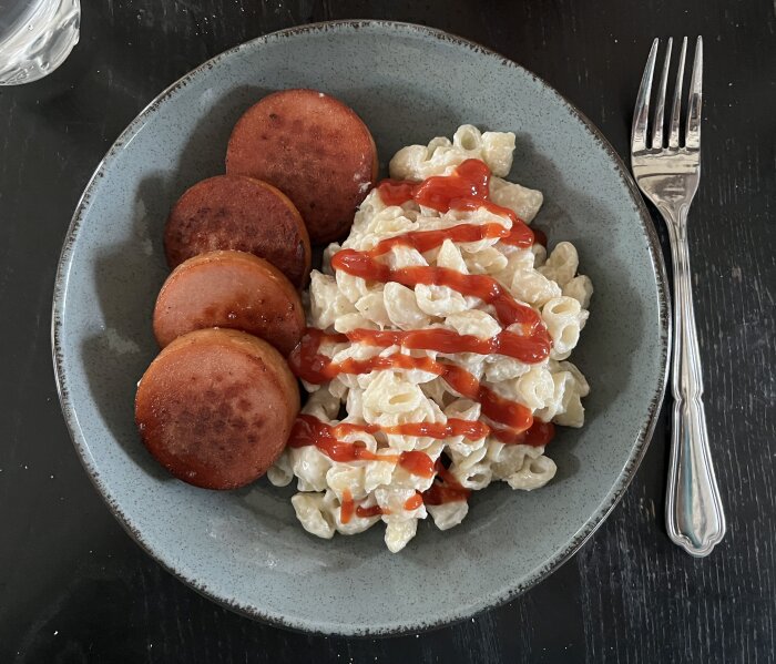 Stuvade makaroner med stekt falukorv och ketchup på en tallrik, serverat med en gaffel på ett bord.