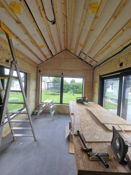 Rum under renovering med isolering och beredskap för köksinstallation. Stegar, byggmaterial och verktyg syns, samt grön utsikt genom fönstren.