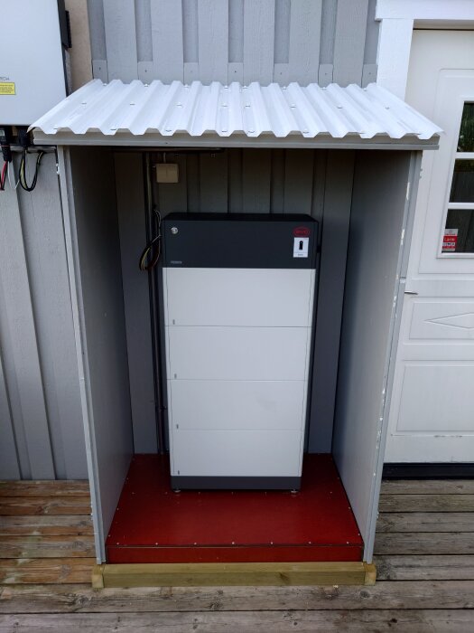 Isolerad batterilåda med frigolit och plywood, med korrugerat plasttak och gångjärn, installerad vid husvägg; designad för frostskydd.