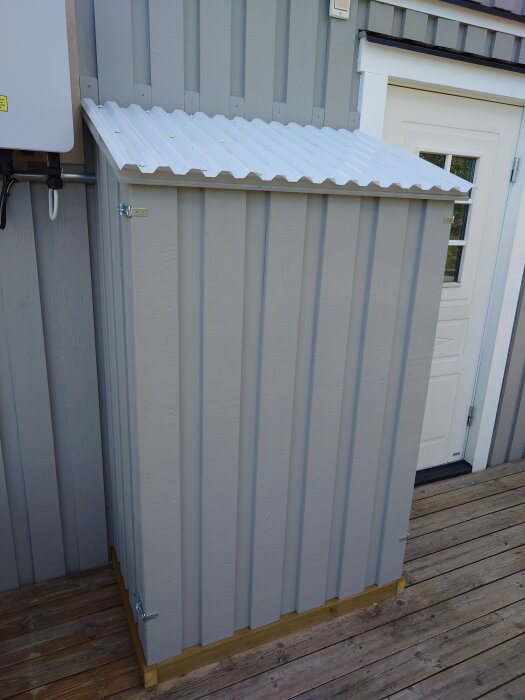 Grå plywoodlåda med korrugerat plasttak, placerad på altan bredvid husvägg.