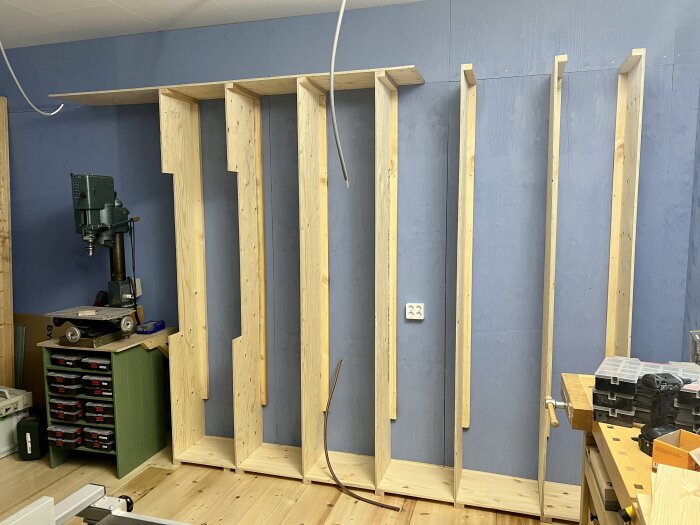 Träramar av plywood uppsatta på en vägg för förvaring i ett snickeriverkstad, med verktyg och en pelarborrmaskin i närheten.