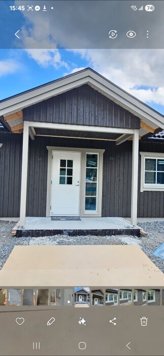 Entré till hus med vit ytterdörr och omålad lockläkt på fasaden framför stenbelagd uppfart, tagen under ett byggprojekt.