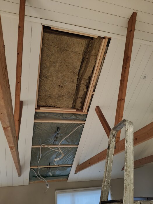 Öppet innertak med synlig isolering och plastfolie, omgiven av träreglar. Stege placerad nära för arbete på höjden.