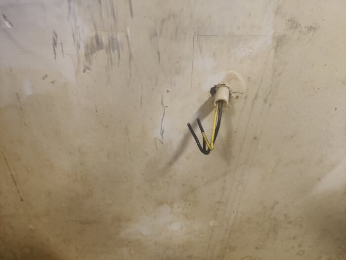 Vägg med tre synliga kablar (två svarta och en gulgrön) som sticker ut från ett runt hål, omgivna av sliten och smutsig yta.