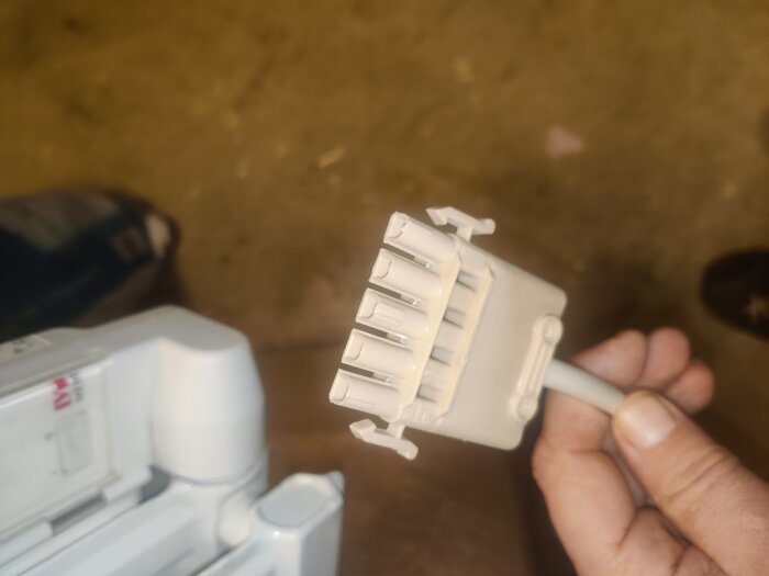 Grå kontakt med flera portar för elektrisk anslutning, hålls i en hand. Text på enheten markerad "LVI".