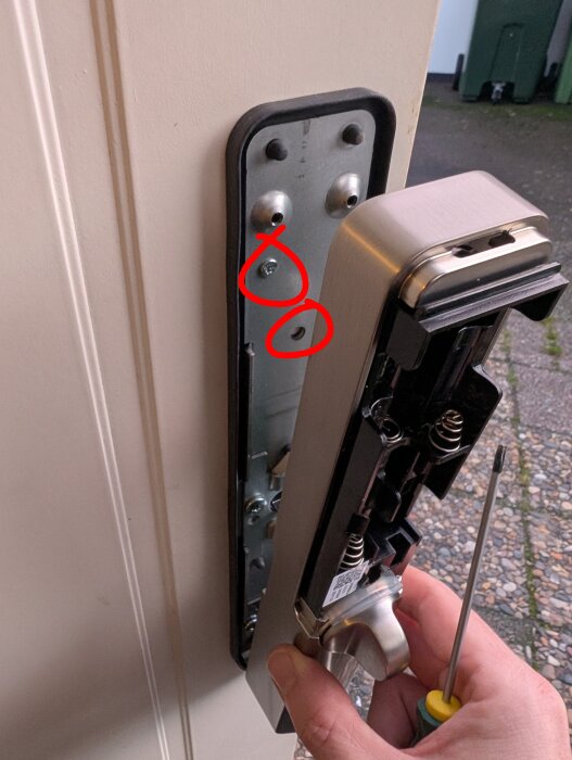 Dörrhandtag isärtaget med synliga skruvar och markerade hål, hand hållandes skruvmejsel visar demontering av låsmekanism.