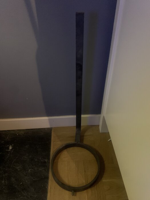 En metallstång med ett stort metallring i ena änden lutad mot en vägg på ett trägolv.