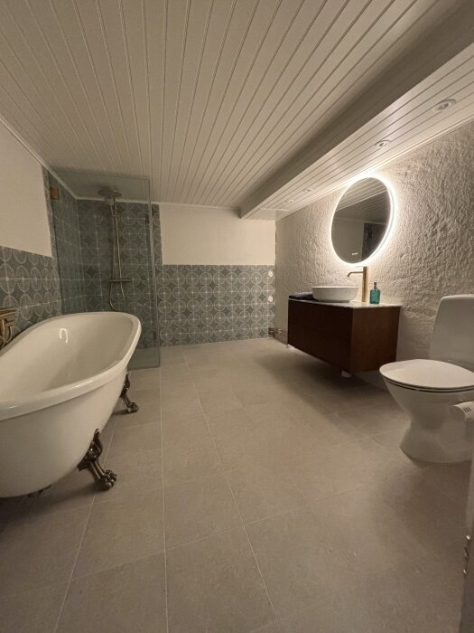 Modernt badrum med fristående badkar, dusch, rund spegel med belysning och toalett på beige kaklat golv.