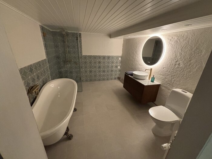 Renoverat badrum med fristående badkar, dusch, rund spegel med belysning, handfat på mörk kommod och toalett.