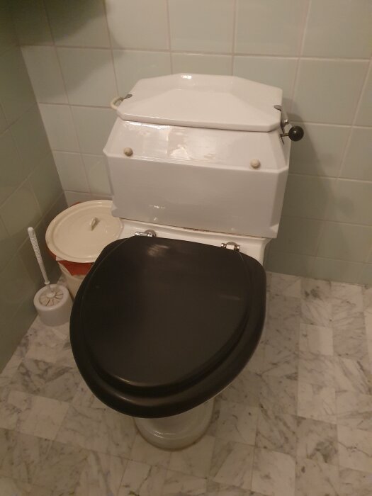 WC med svart sits på mönstrat golv, vit cistern och borste bredvid.