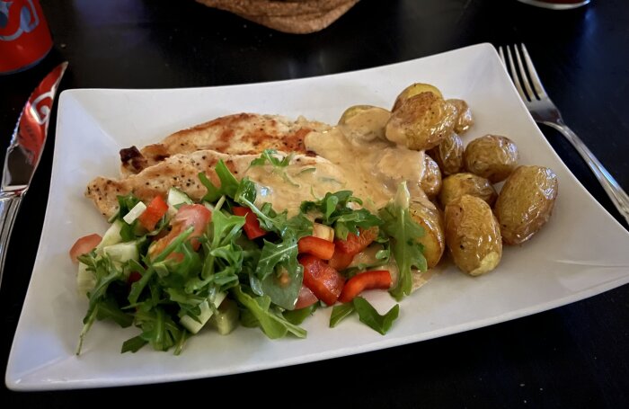 Stekt kycklingfilé, ugnstekt småpotatis, grönsallad och sås serverat på en vit tallrik.