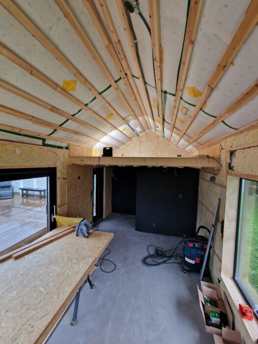 Renovering pågår, med plywood som fönsterfoder och exponerade takbjälkar. Byggmaterial och verktyg synliga i rummet.